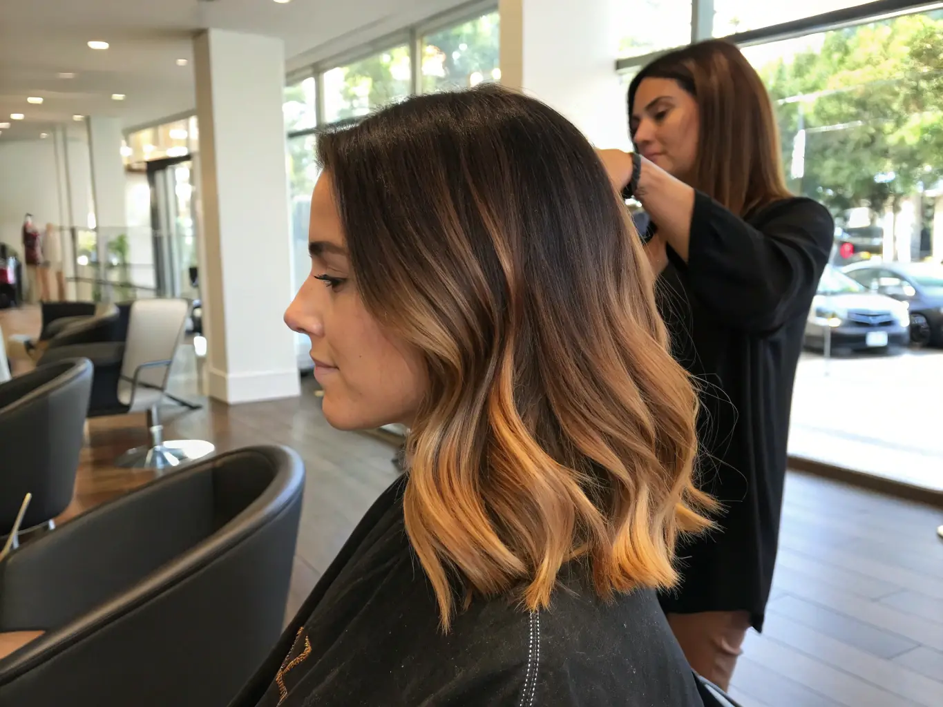 A close-up shot of a woman's hair showcasing a perfectly blended balayage, with soft, natural-looking highlights that transition seamlessly from dark roots to lighter ends, styled in loose waves to emphasize the color dimension, in a salon setting.
